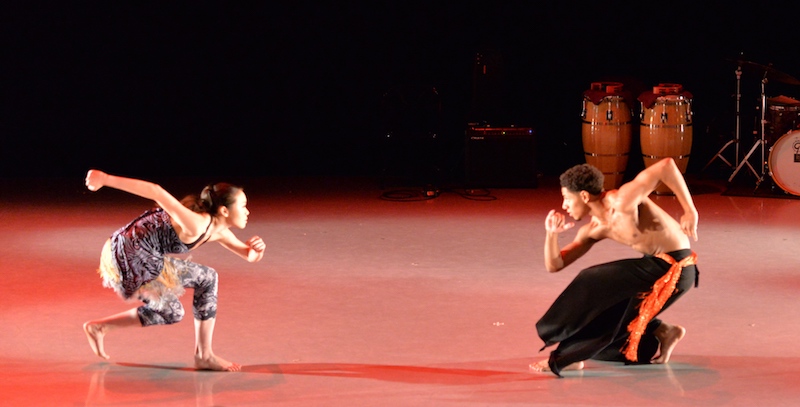 Two dancers crouch and look toward one another. The stage is brightly lit and there are percussion instruments in the background.